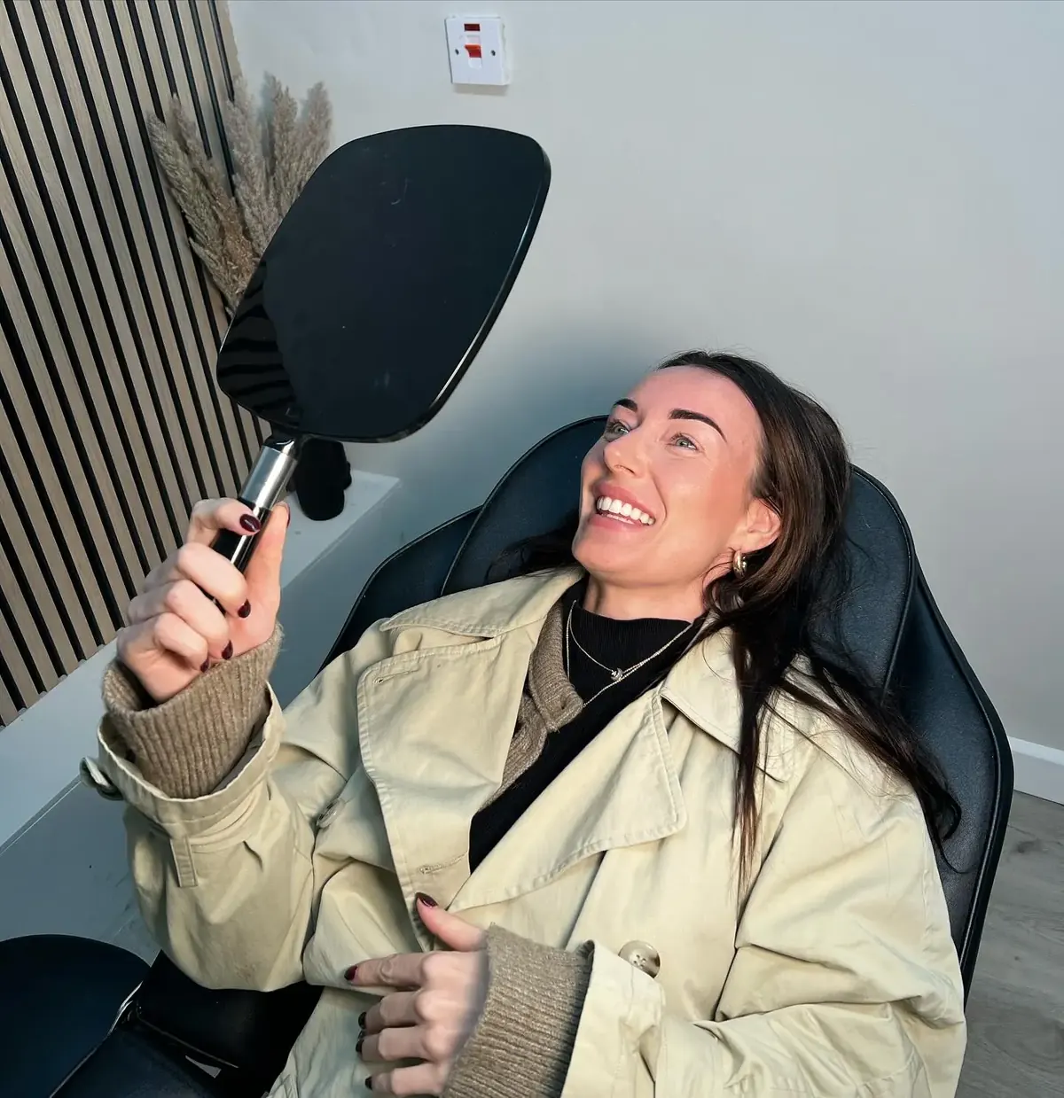 Client smiling while looking at her freshly treated eyebrows in a handheld mirror after a permanent makeup appointment.